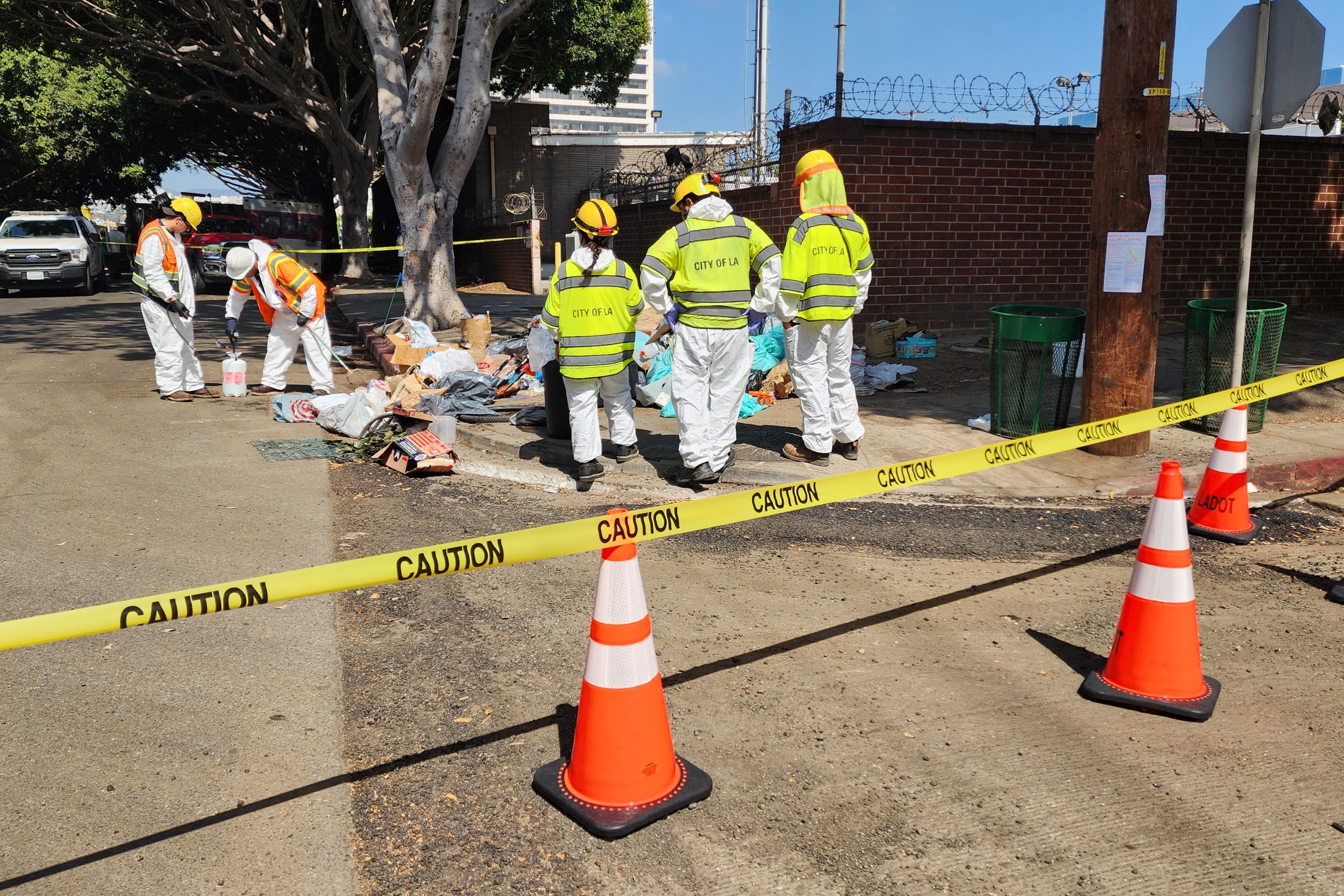 A line of caution tape separates the photographer from three sanitation workers standing in front of a pile of discarded trash and other belongings that were abandoned.