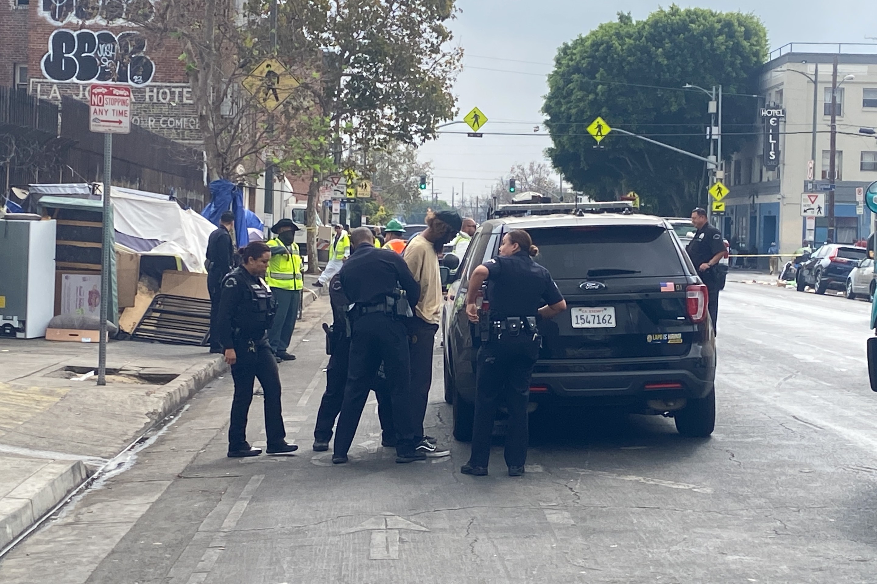 L.A. TACO Staff Investigative Reporter Lexis-Olivier Ray, is handcuffed while filming a sanitation "sweep." Officers are surrounding Ray and two officers are holding his arms and placing cuffs on him